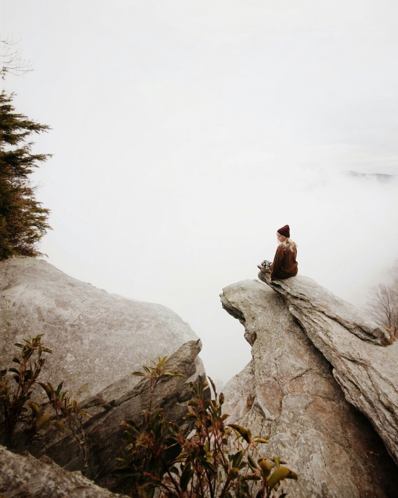A woman sits alone on a rocky cliff edge surrounded by fog and mist, looking into the distance — symbolising solitude, reflection, and the inner landscape of grief.